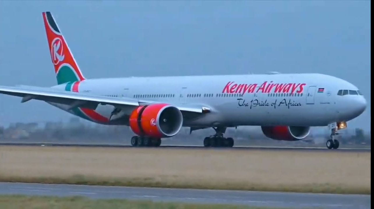 White Kenya Airways Boeing 777 with red engines and green-red tail taxiing on a rainy runway, displaying “The Pride of Africa” slogan.